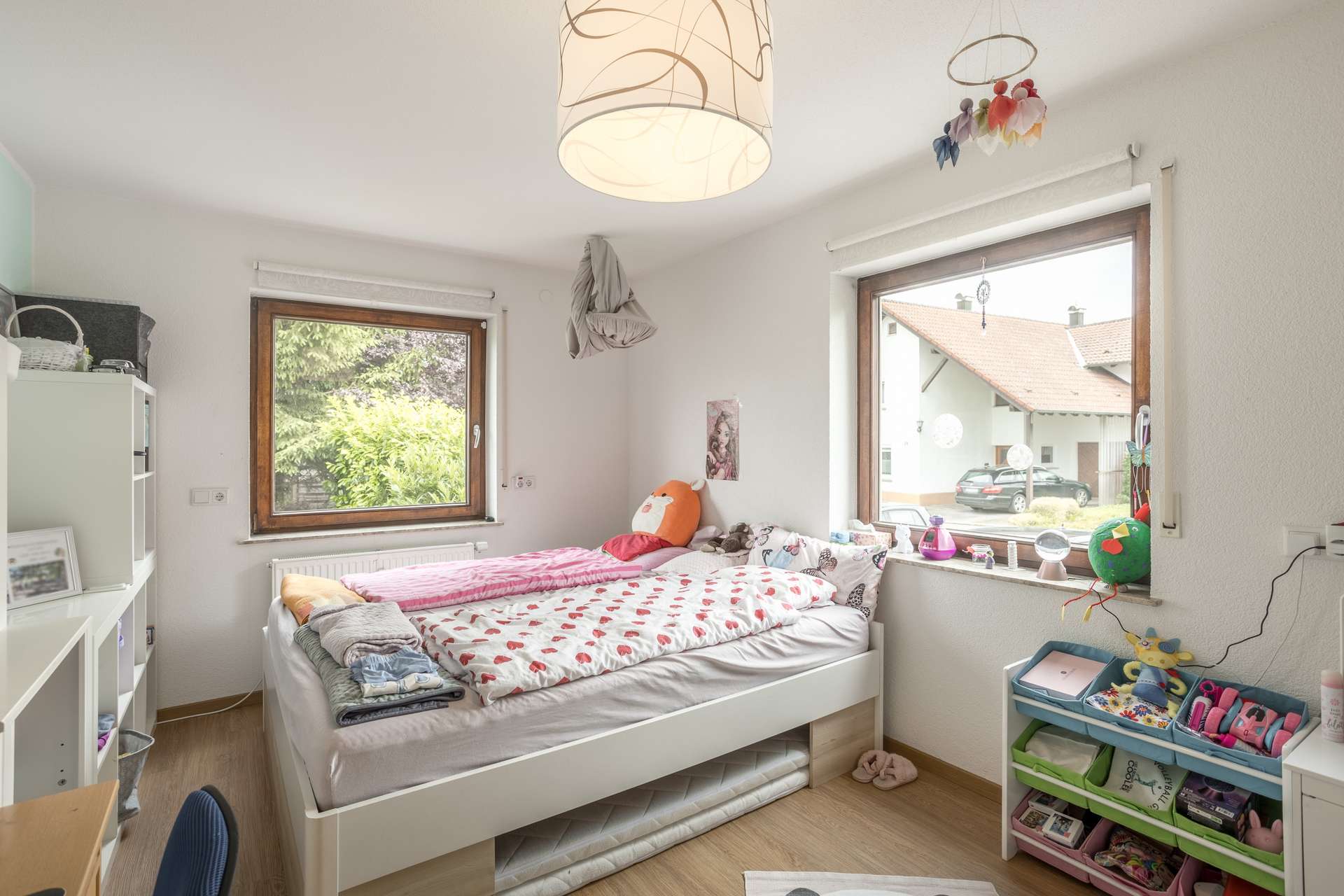 EG Ein Kinderzimmer mit einem weißen Bett mit Bettwäsche mit Herzmuster, zwei Fenstern und bunten Aufbewahrungsbehältern.