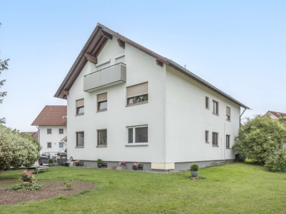 Ein dreistöckiges weißes Wohnhaus mit Balkon und Satteldach, das in einem begrünten Garten steht.
