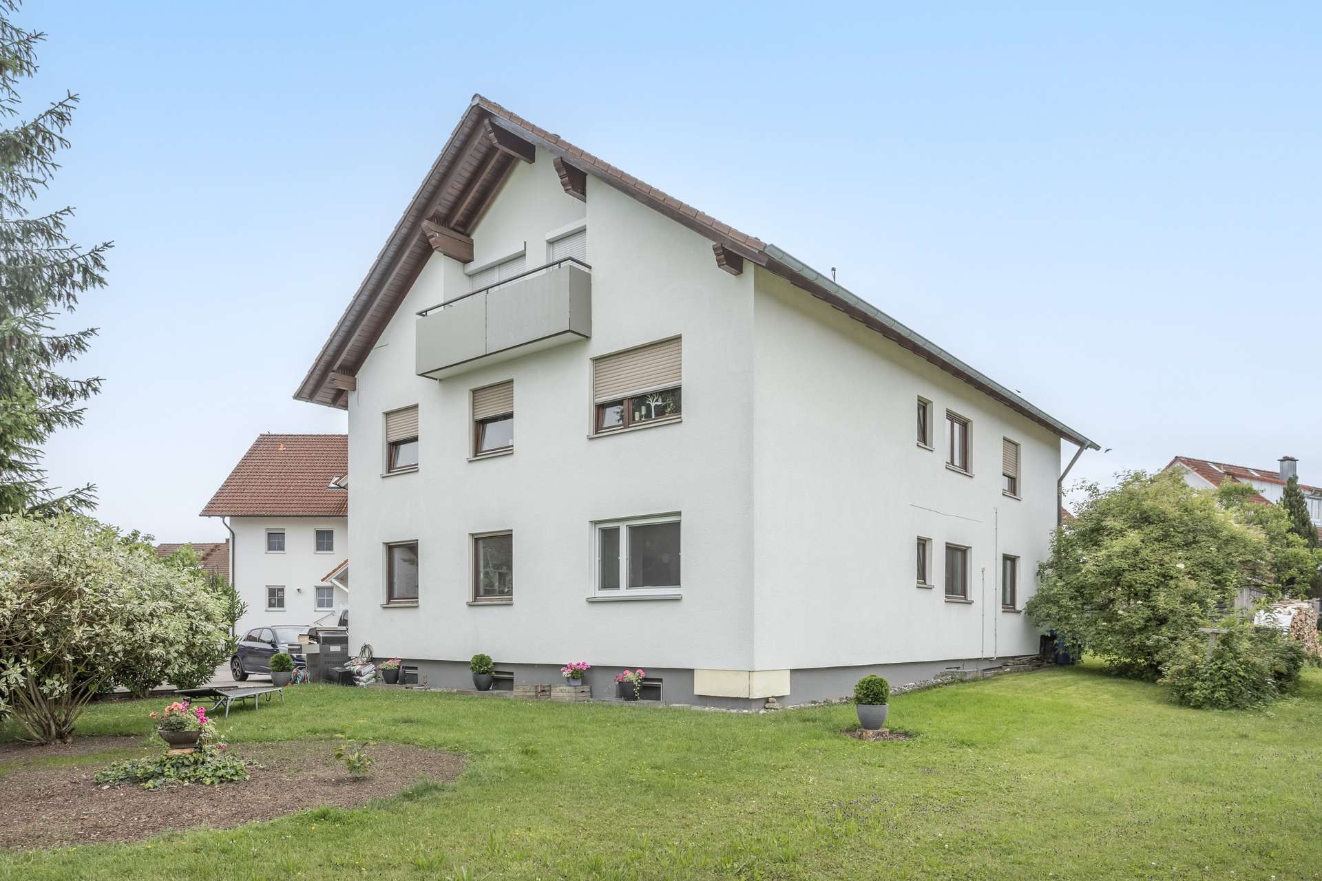 Außenansicht Ein dreistöckiges weißes Wohnhaus mit Balkon und Satteldach, das in einem begrünten Garten steht.