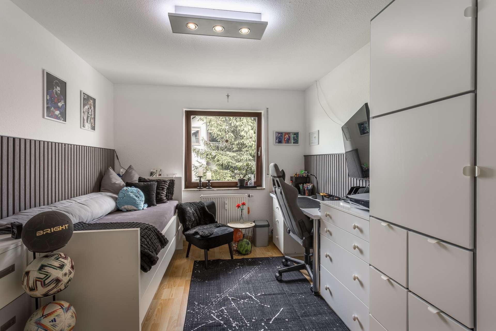 1.OG Ein Schlafzimmer mit einem Einzelbett, einem Schreibtisch mit einem Gaming-Stuhl, einer Kommode und einem Fenster mit Blick auf Bäume.