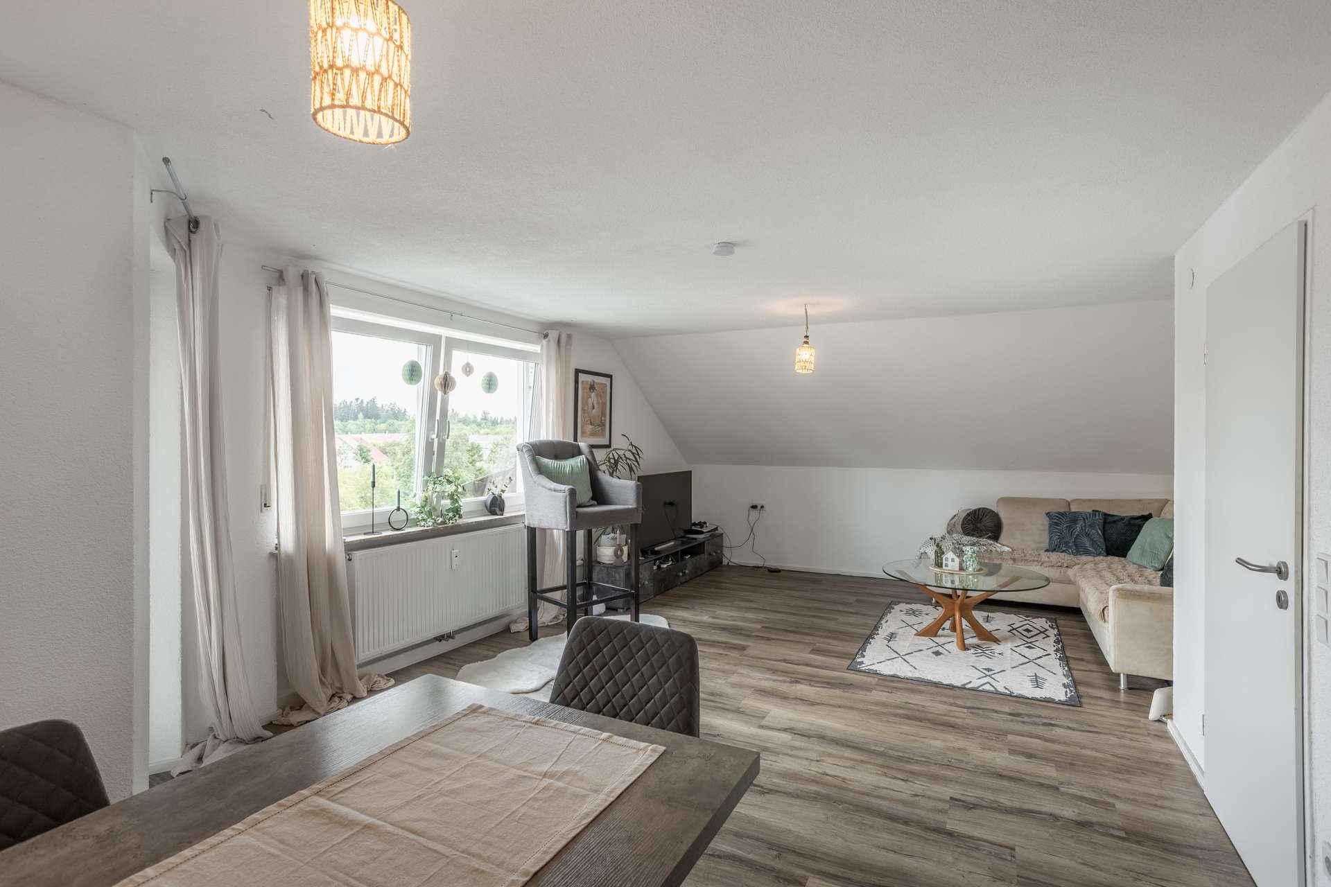 DG Ein offen gestaltetes Wohnzimmer mit einem Esstisch im Vordergrund, einem Ecksofa mit einem Couchtisch aus Glas und einem Fenster mit Aussicht.