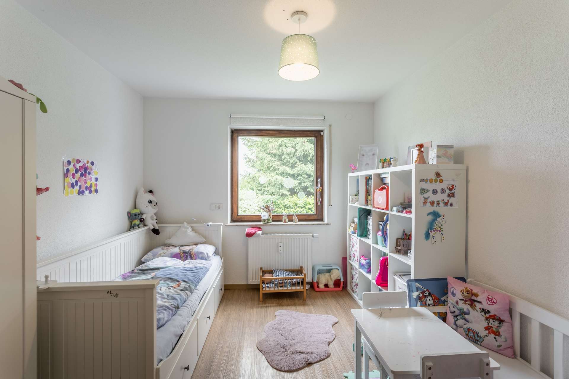 EG Ein Kinderzimmer mit einem Einzelbett, einem Fenster, einem weißen Regal voller Spielsachen und einem kleinen Tisch mit Stühlen.