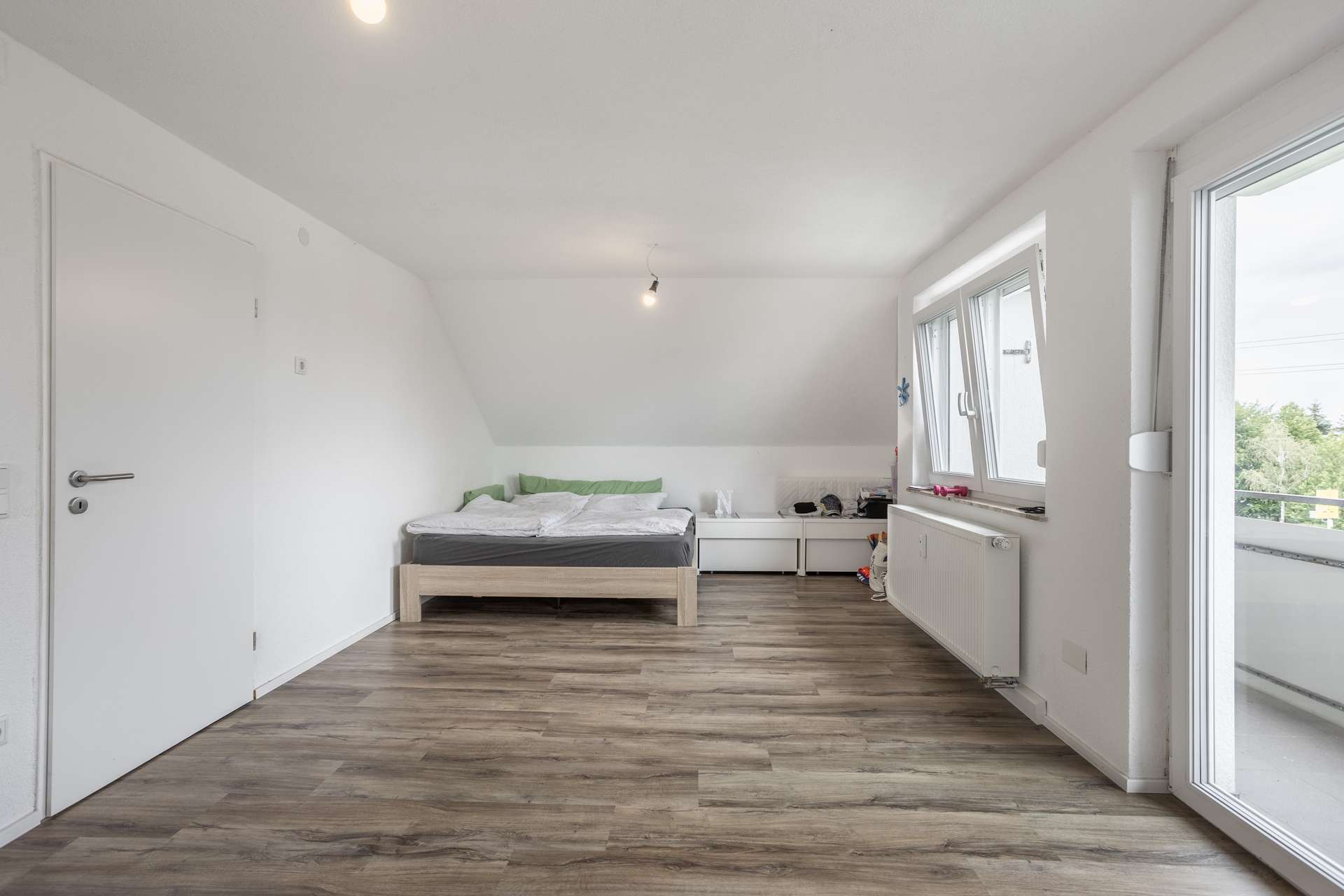 DG Ein Schlafzimmer mit einem niedrigen Bettgestell aus Holz, weißen Wänden, einem Bodenbelag in Holzoptik und einer Glastür, die auf einen Balkon führt.