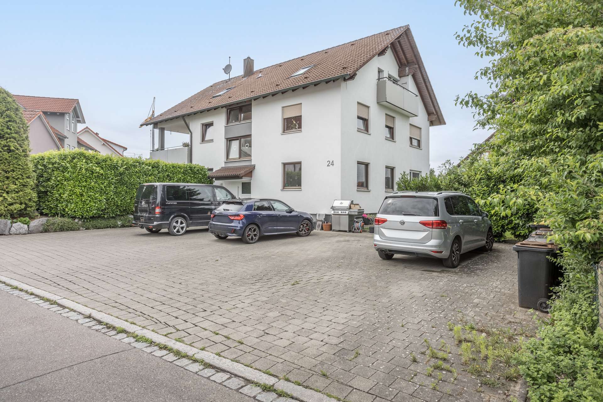 Außenansicht Ein mehrstöckiges Wohnhaus mit weißer Fassade und Ziegeldach, vor dem sich ein gepflasterter Parkplatz mit drei geparkten Autos befindet.
