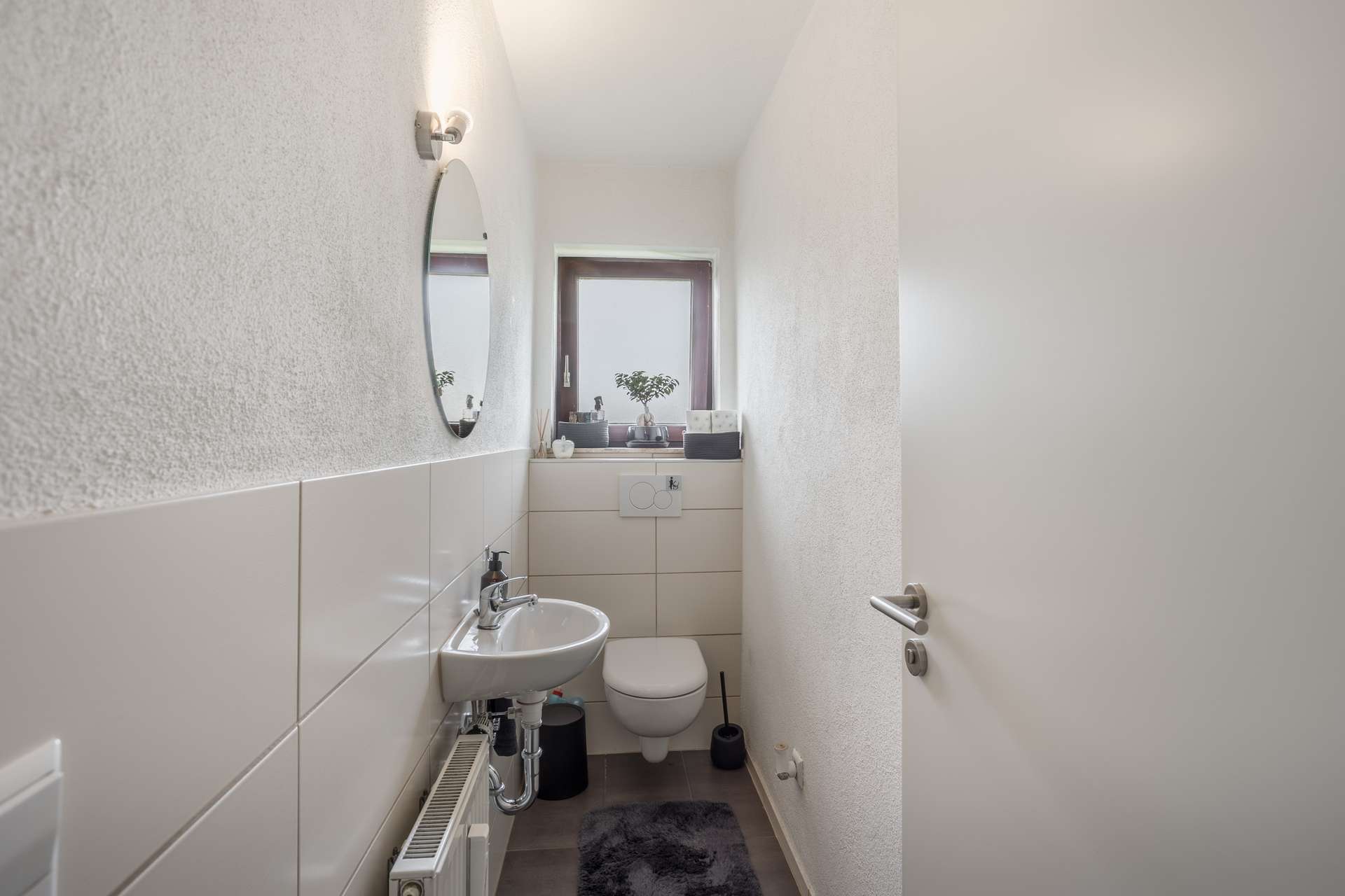 1.OG Ein kleines Badezimmer mit einem an der Wand montierten Waschbecken, einer Toilette, einem runden Spiegel und einem Fenster über der Toilette.