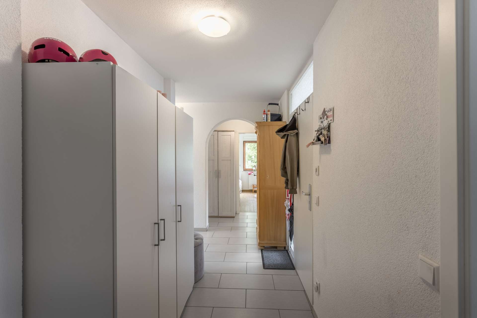EG Ein Flur mit weißen Kleiderschränken auf der linken Seite und einer Garderobe auf der rechten Seite, der durch einen Torbogen in einen sonnendurchfluteten Raum führt.