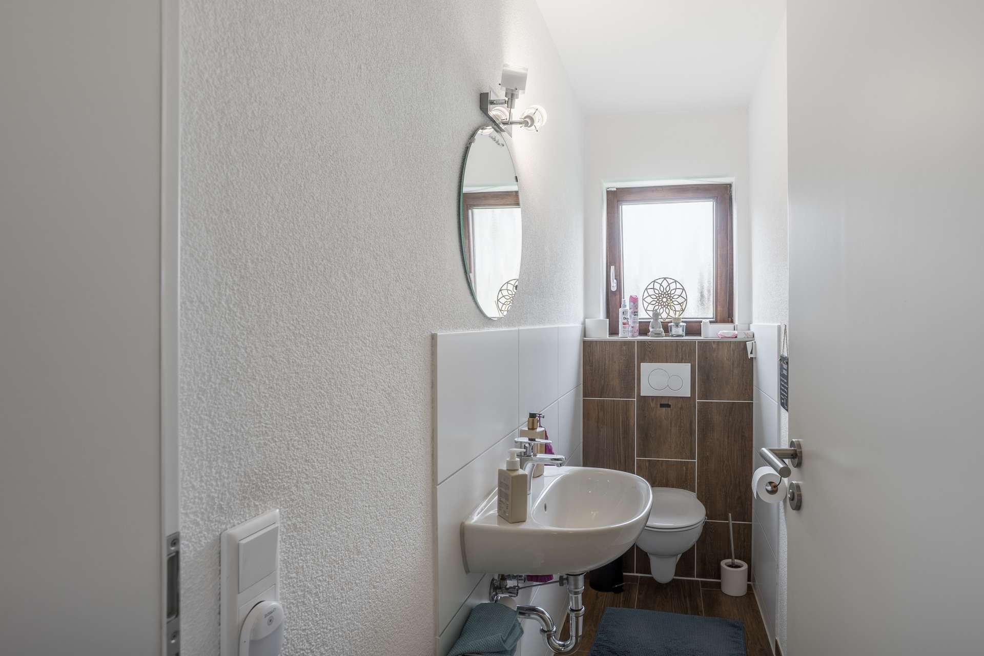 EG Kleines Badezimmer mit einem wandmontierten Waschbecken, einer Toilette, einem runden Spiegel und einem schmalen Fenster.