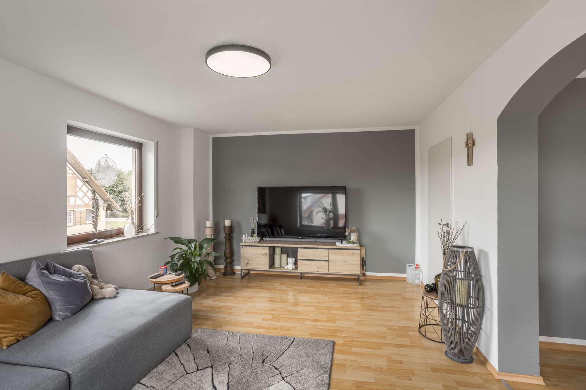1.OG Ein Wohnzimmer mit einem grauen Sofa, einem TV-Schrank in Holzoptik, auf dem ein Fernseher steht, und einer Akzentwand vor hellem Holzfußboden.