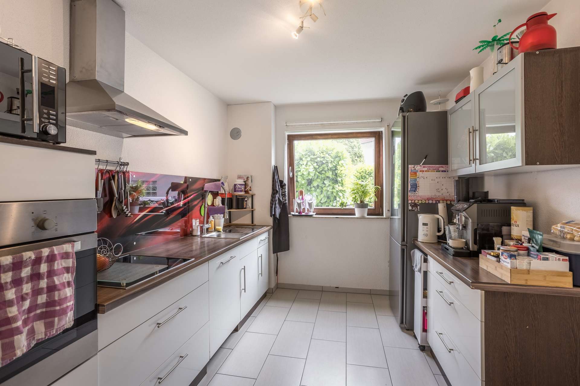 EG Eine Küche im Galley-Stil mit weißen Schränken, einer Arbeitsplatte aus dunklem Holz, einem Edelstahl-Backofen, einem Kühlschrank und einem Fenster mit Blick ins Grüne.