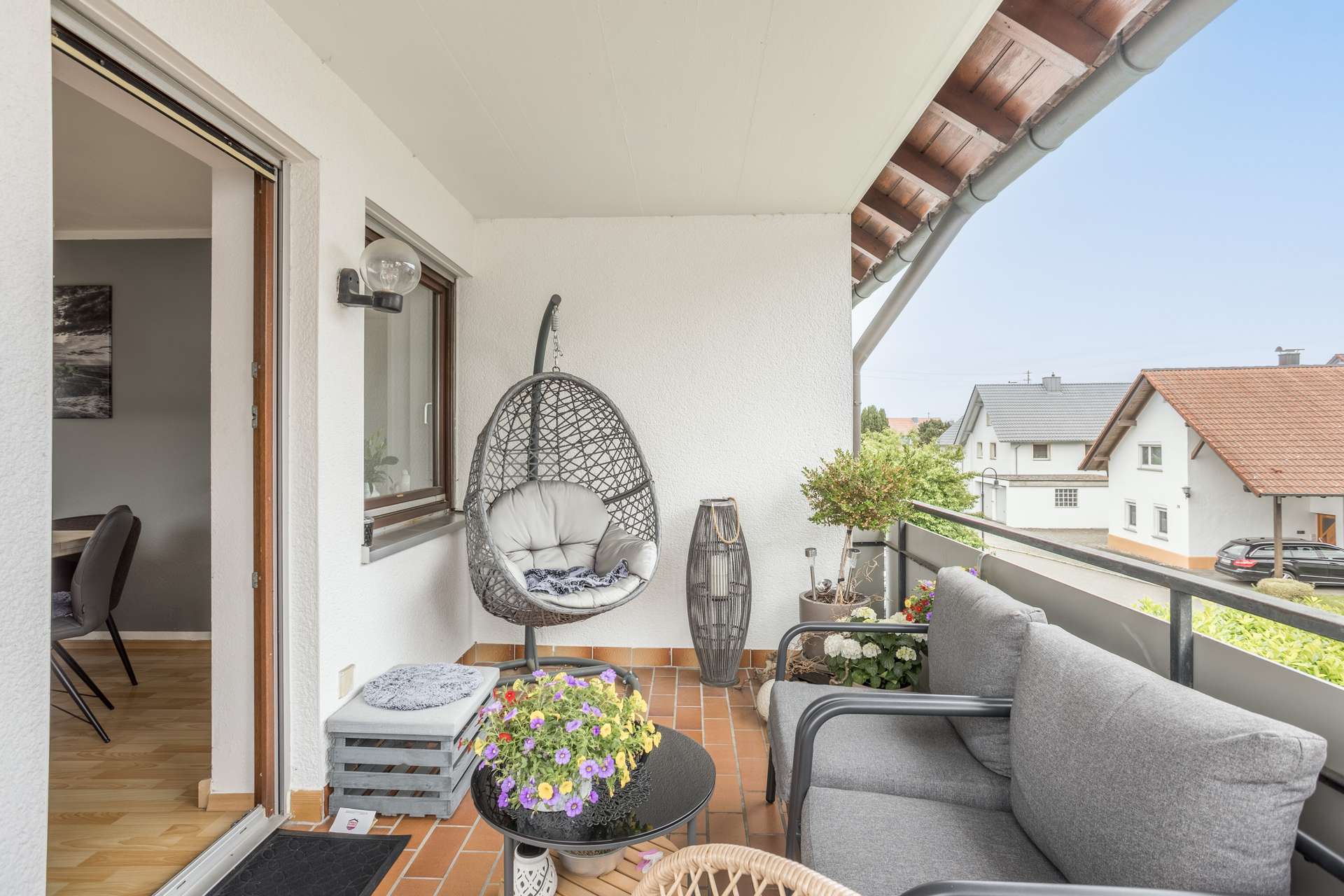 1.OG Ein Balkon mit einem hängenden Eiersessel, einem grauen Sofa, einem kleinen runden Tisch mit Topfblumen und Blick auf die Nachbarhäuser.