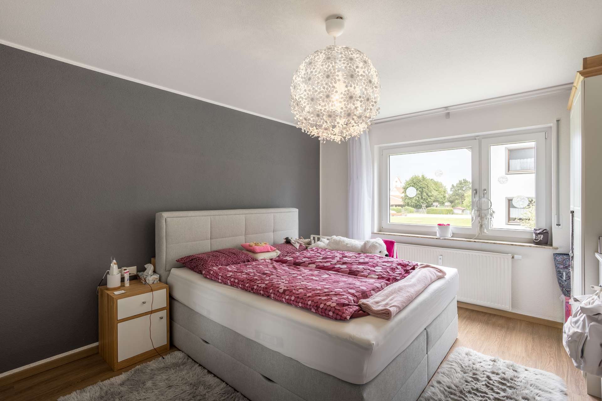 EG Ein Schlafzimmer mit einem grauen Polsterbett, einer burgunderroten Bettdecke mit Muster und einer großen weißen Pendelleuchte mit Blumenmuster.