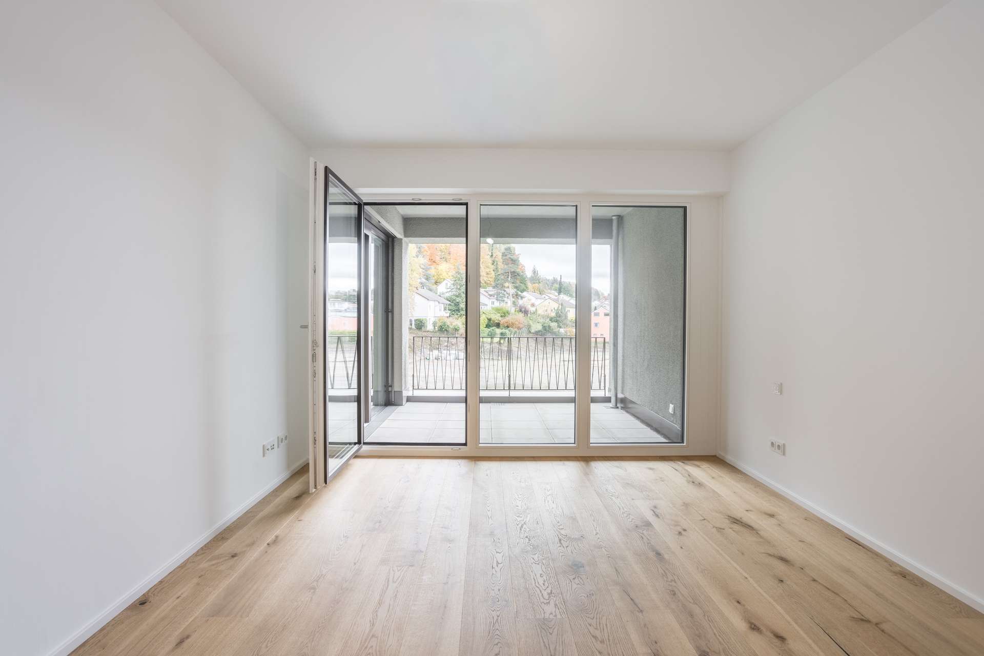 Ein leerer Raum mit weißen Wänden, hellem Holzboden und großen Glastüren, die auf einen Balkon mit Blick auf Bäume und Häuser führen. Ein leerer Raum mit weißen Wänden, hellem Holzboden und großen Glastüren, die auf einen Balkon mit Blick auf Bäume und Häuser führen.