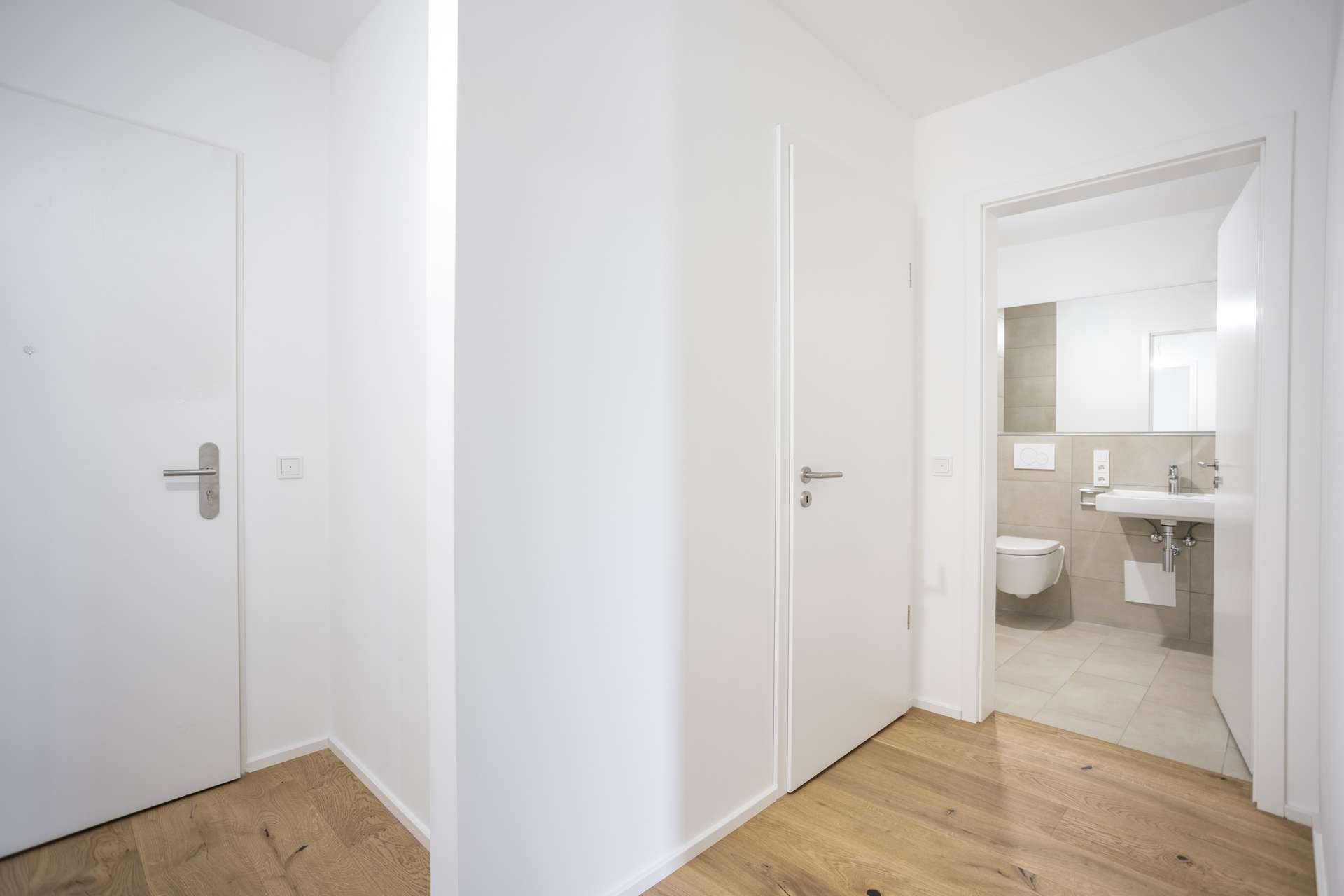 Blick auf einen Flur mit weißen Türen und hellem Holzboden, der in ein Badezimmer mit einem wandmontierten WC und einem Waschbecken führt. Blick auf einen Flur mit weißen Türen und hellem Holzboden, der in ein Badezimmer mit einem wandmontierten WC und einem Waschbecken führt.