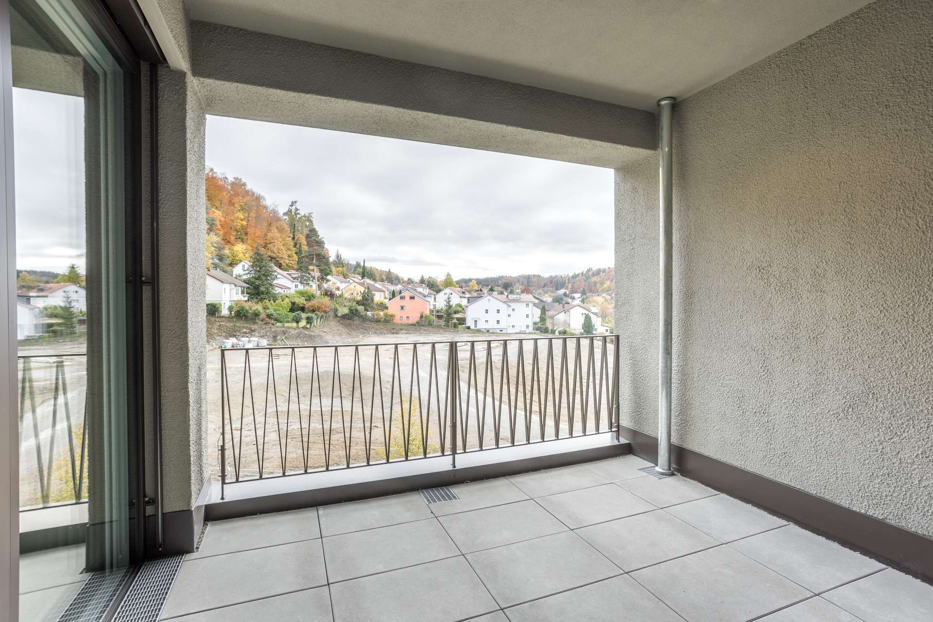 Ein überdachter Balkon mit Metallgeländer, von dem aus man auf ein unbebautes Grundstück und ein am Hang gelegenes Wohnviertel mit Häusern blickt. Ein überdachter Balkon mit Metallgeländer, von dem aus man auf ein unbebautes Grundstück und ein am Hang gelegenes Wohnviertel mit Häusern blickt.