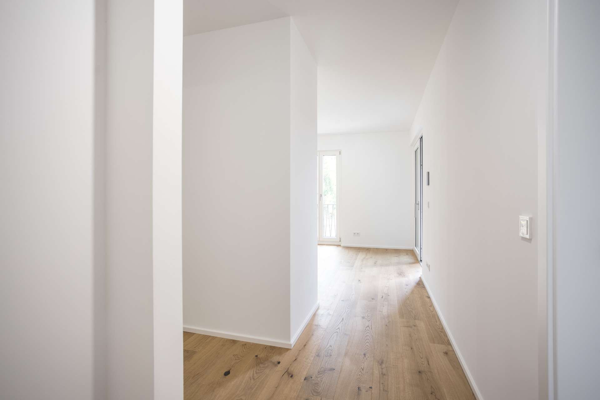Ein leerer, weißer Flur mit hellem Holzboden und Blick auf einen hellen Raum mit einer gläsernen Balkontür am Ende. Ein leerer, weißer Flur mit hellem Holzboden und Blick auf einen hellen Raum mit einer gläsernen Balkontür am Ende.