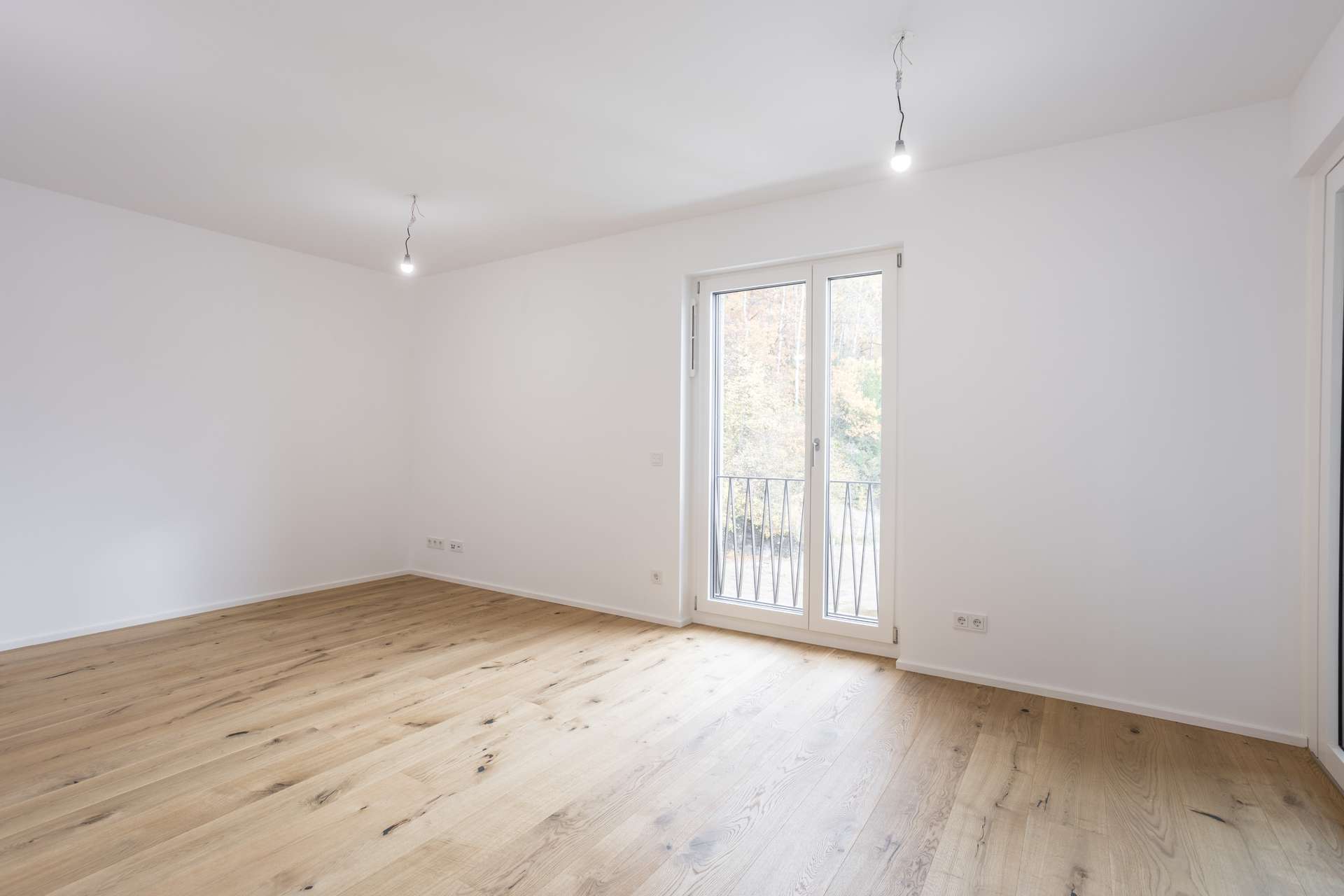Ein leerer Raum mit weißen Wänden, Holzboden und einer gläsernen Balkontür, durch die man auf Bäume blickt. Ein leerer Raum mit weißen Wänden, Holzboden und einer gläsernen Balkontür, durch die man auf Bäume blickt.